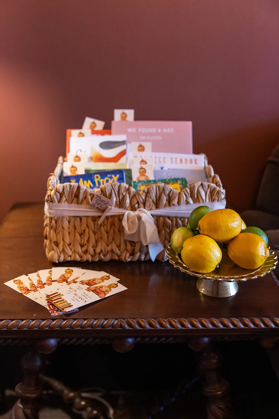A woven basket of picture books tied with ribbon on an antique table, beside a bowl of lemons and a stack of party invitations