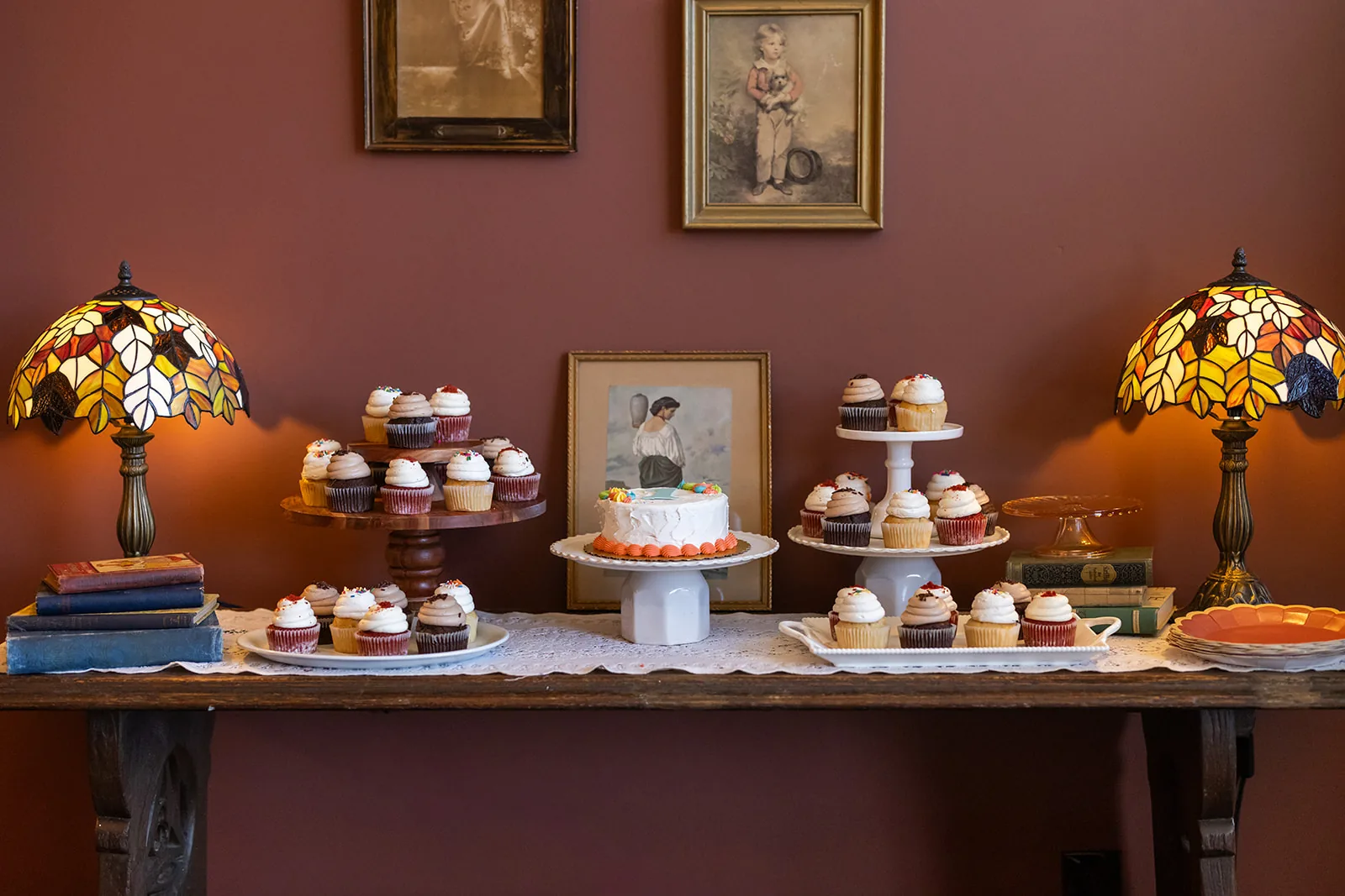 A dessert table of tiered cupcake stands and a decorated cake, flanked by stained glass lamps and framed vintage artwork