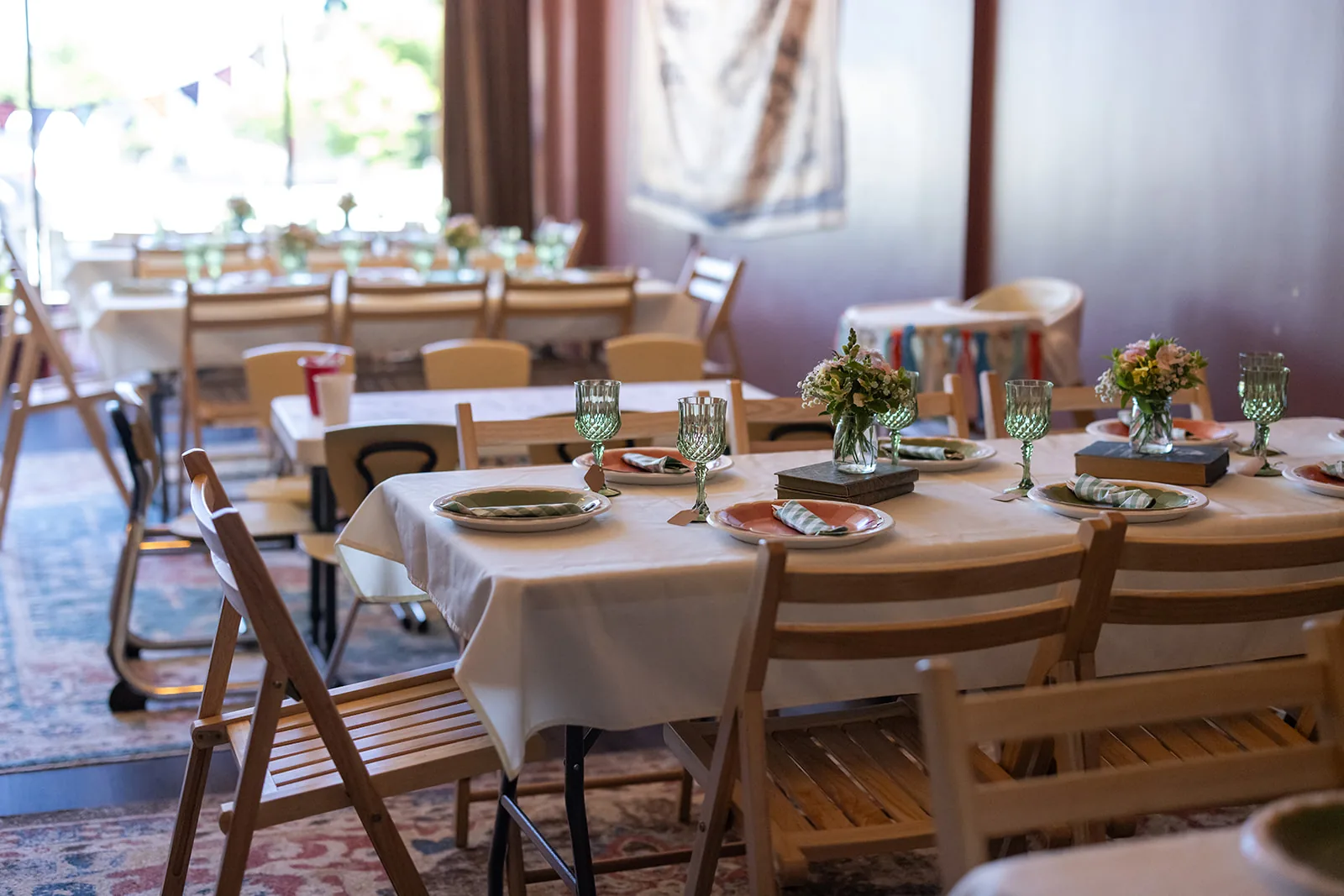 Long tables set for a birthday party in the event space at The Wandering Lantern, with white linens, green glassware, and flowers on stacked vintage books