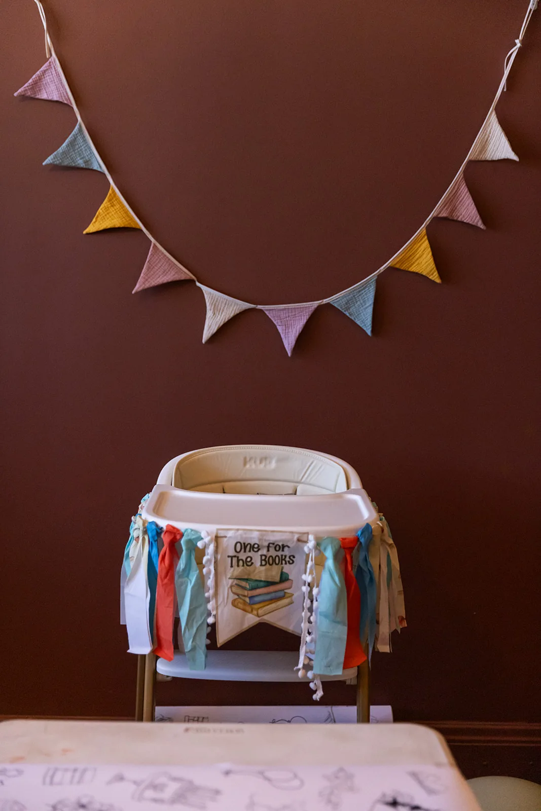 A high chair decorated with a One for the Books tassel banner beneath a string of pastel bunting