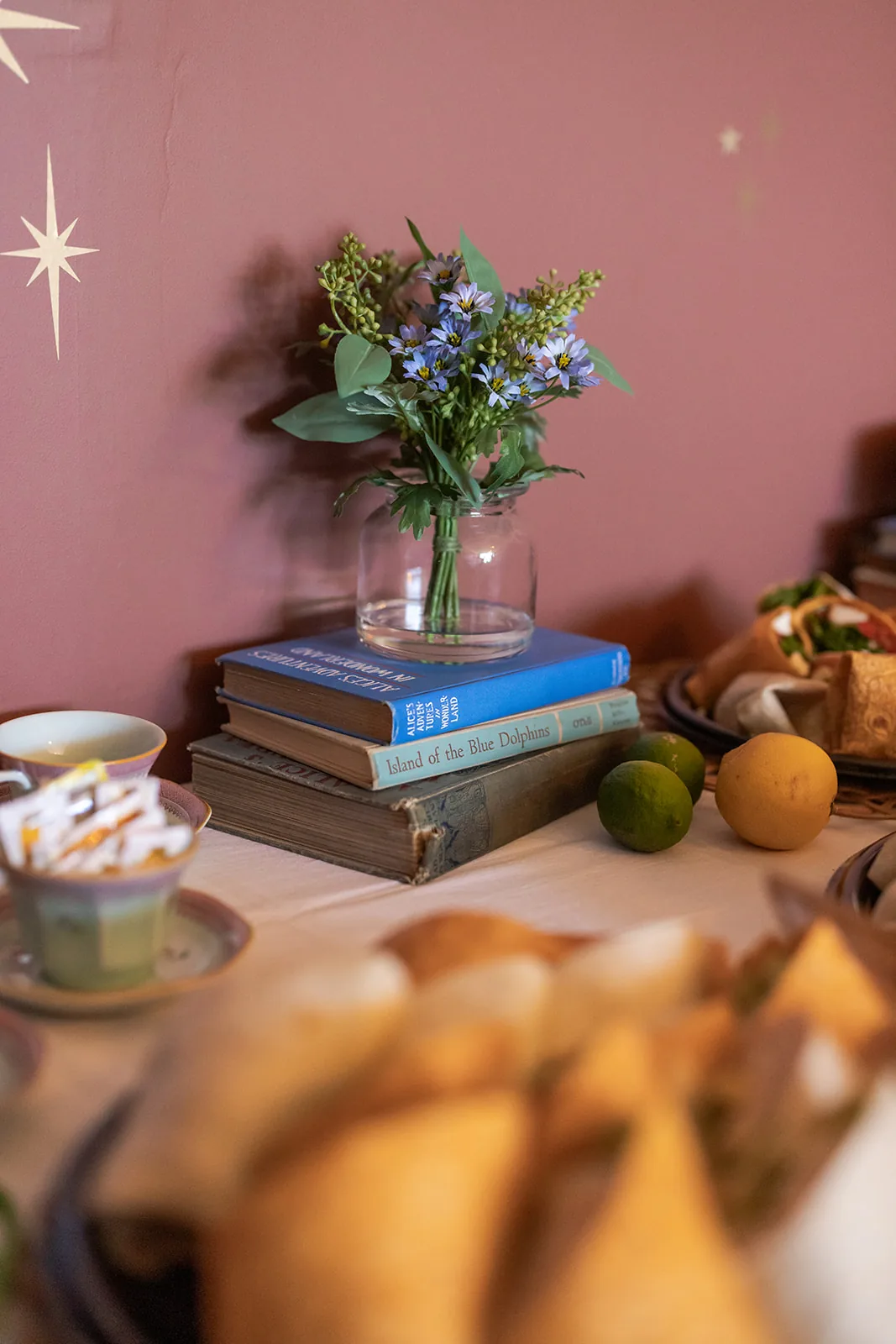 A jar of wildflowers resting on a stack of vintage hardcover books against a deep rose wall with small star decals
