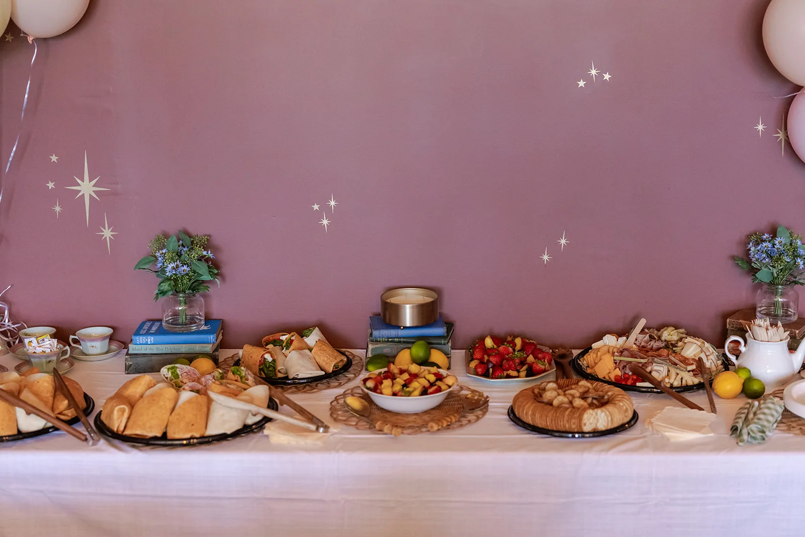 A long grazing table spread with charcuterie, pastries, fruit, and desserts, with small floral arrangements on stacked books