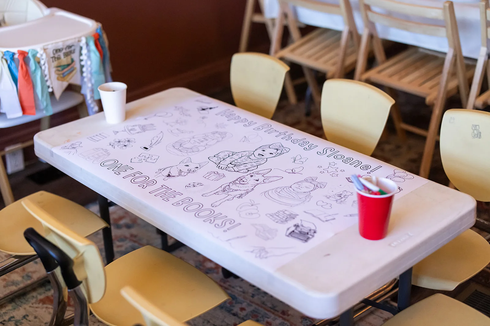 A kids activity table covered with a printed coloring sheet and crayons, surrounded by child sized chairs