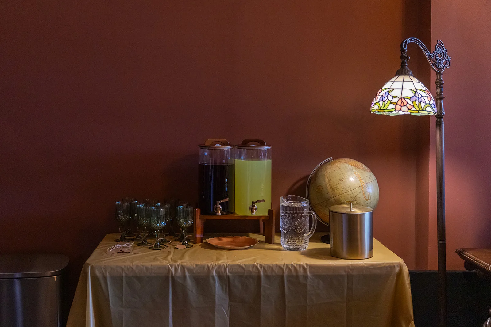 A drink station with two glass beverage dispensers of lemonade, stacked glassware, and an antique globe beside a stained glass floor lamp
