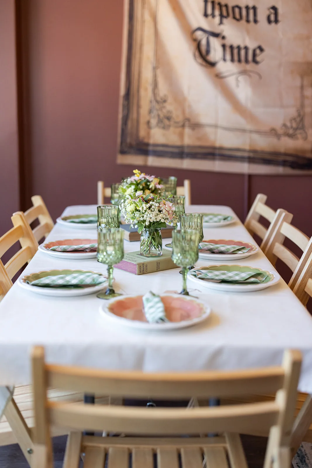 A set party table beneath an Once Upon a Time banner, with gingham napkins, scalloped plates, and a jar of wildflowers on vintage books