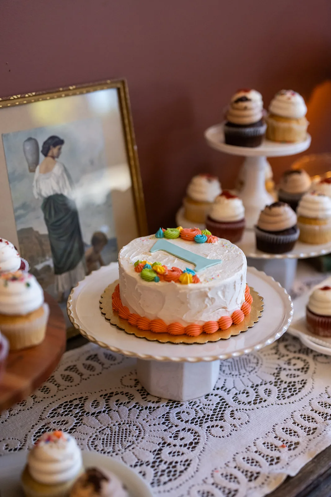 A small white smash cake on a footed stand surrounded by frosted cupcakes and a framed vintage illustration
